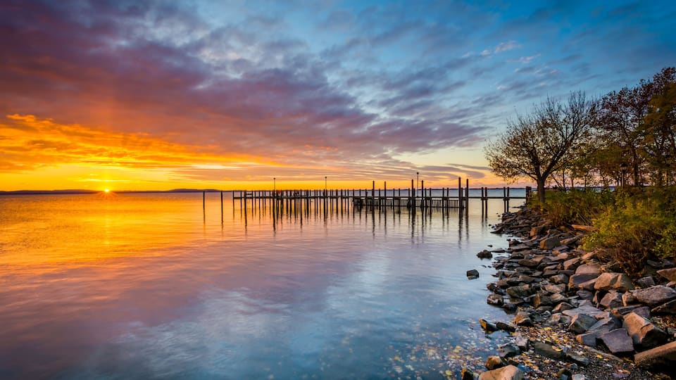 A sunset over a calm lake with a wooden pier extending into the water, surrounded by rocks and trees.