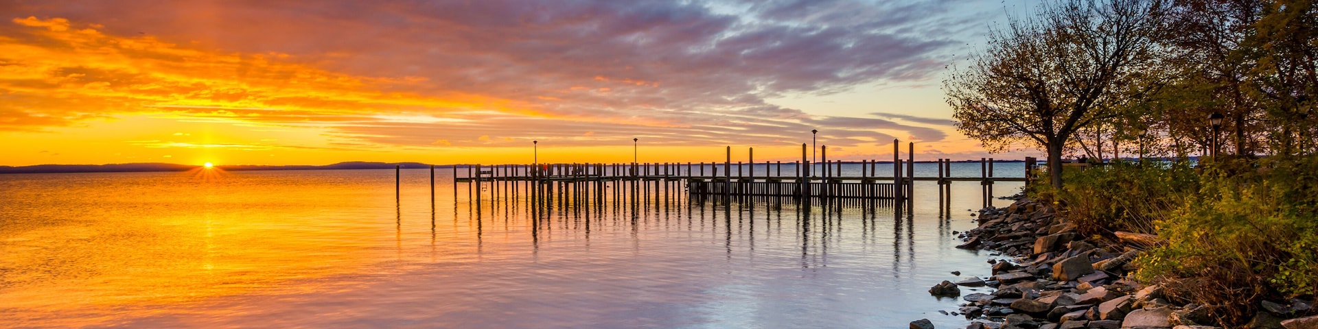 Un atardecer sobre un lago tranquilo con un muelle de madera que se adentra en el agua, rodeado de rocas y árboles.