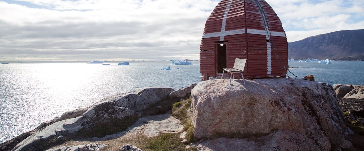 A red wooden structure with a bench, situated on a rocky shore with a view of the ocean and icebergs.