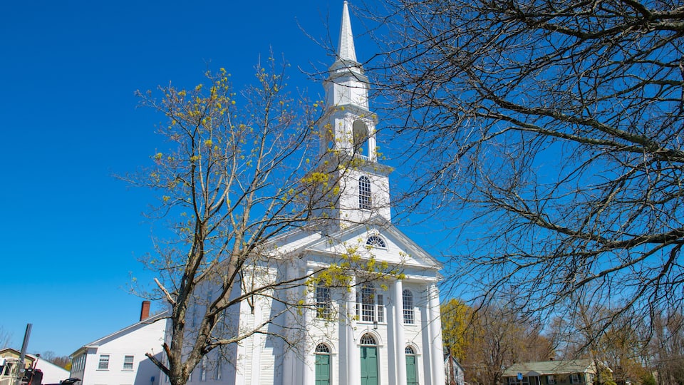 A white church with a steeple and green doors, surrounded by trees and a clear blue sky.