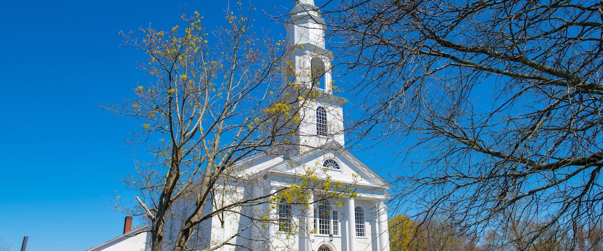 A white church with a steeple and green doors, surrounded by trees and a clear blue sky.