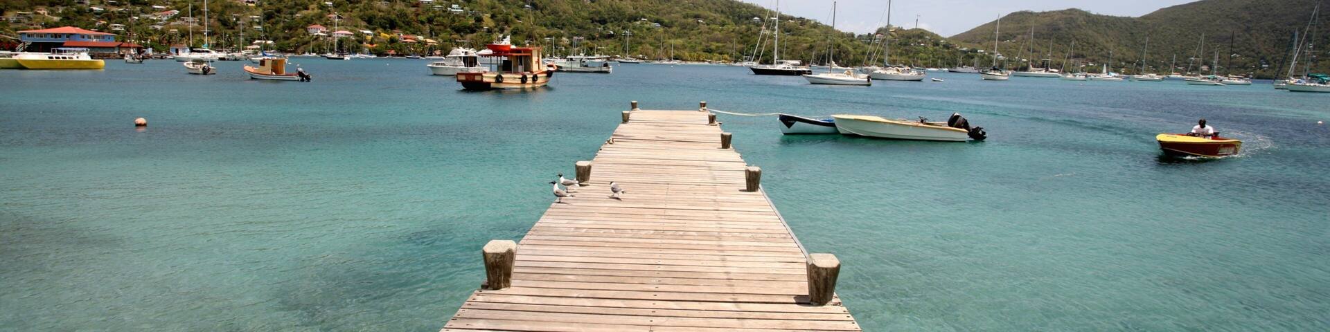 A wooden pier extending into turquoise waters with boats and a hilly backdrop.