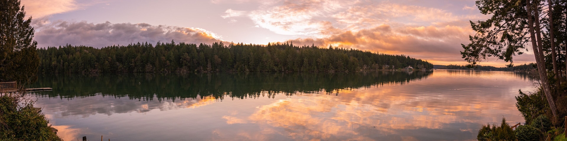 A calm lake surrounded by dense forest, with a wooden dock on the left and a clear reflection of the sky and clouds on the water.