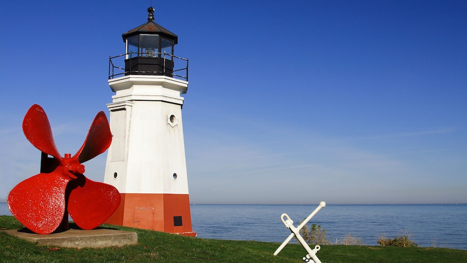 A lighthouse with a red and white color scheme, a large red anchor sculpture, and a white anchor sculpture on a grassy area by the water.