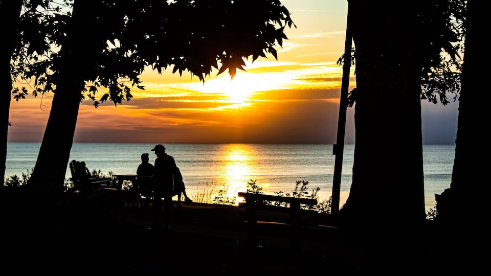 A silhouette of people sitting on a bench at sunset by the water, with trees framing the scene.