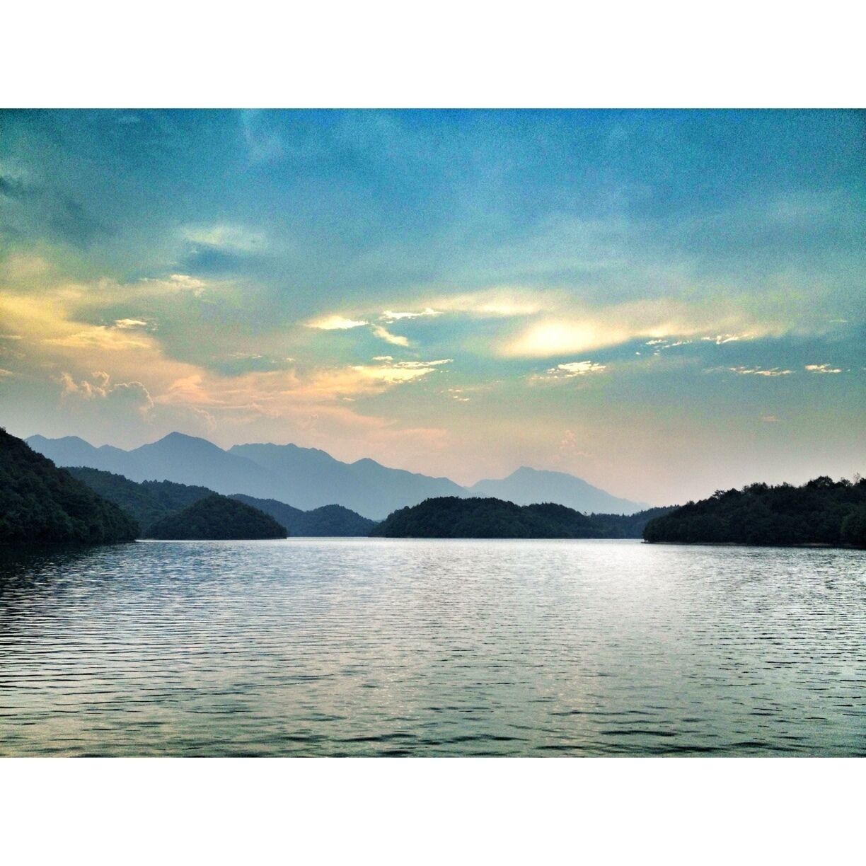 A calm lake surrounded by forested hills under a partly cloudy sky.