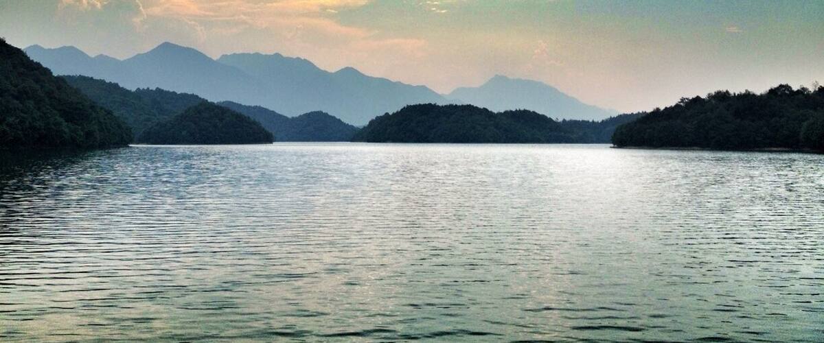 A calm lake surrounded by forested hills under a partly cloudy sky.
