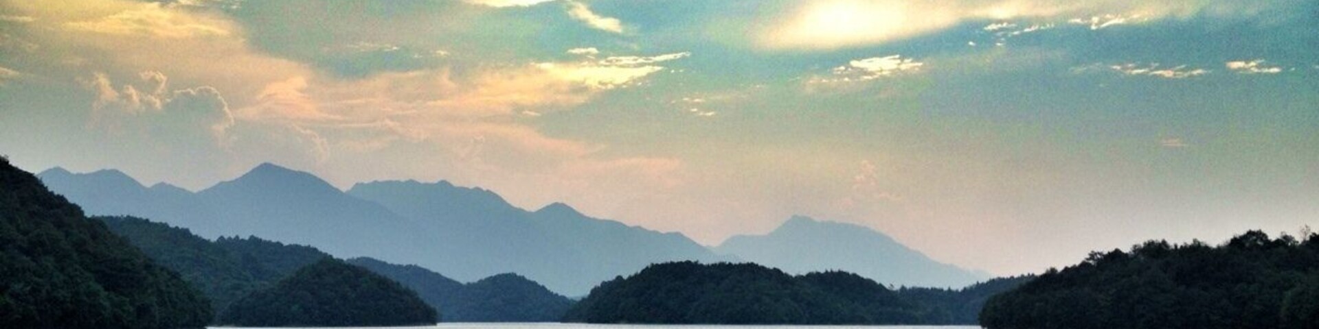 A calm lake surrounded by forested hills under a partly cloudy sky.
