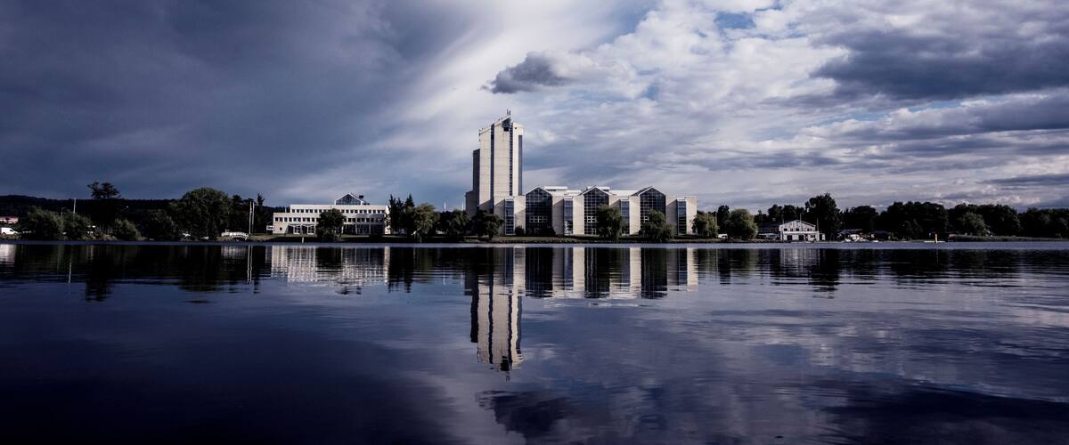 A calm lake with a reflection of buildings and a cloudy sky.