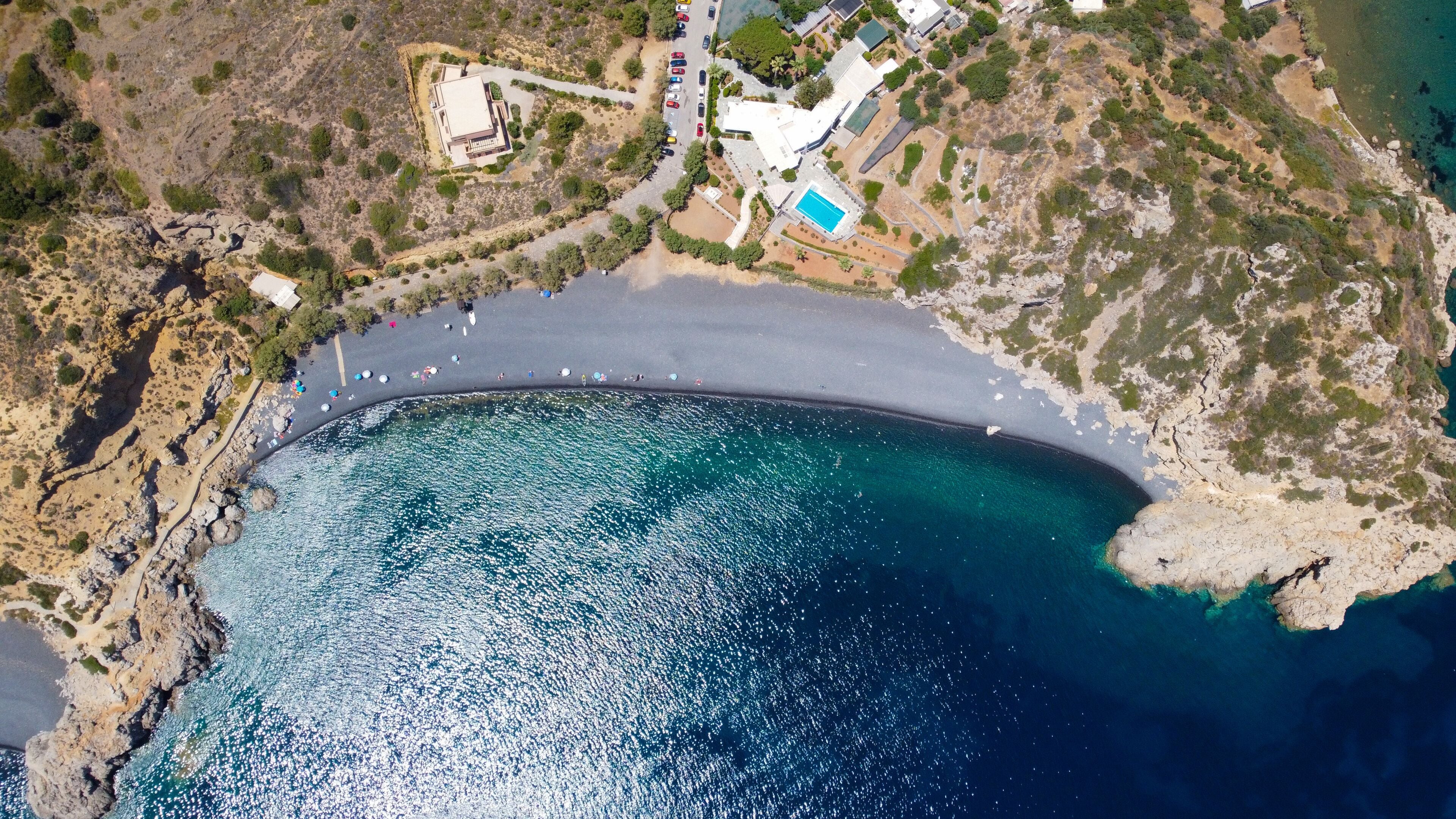 A coastal area with a clear blue sea, rocky terrain, and a few buildings.