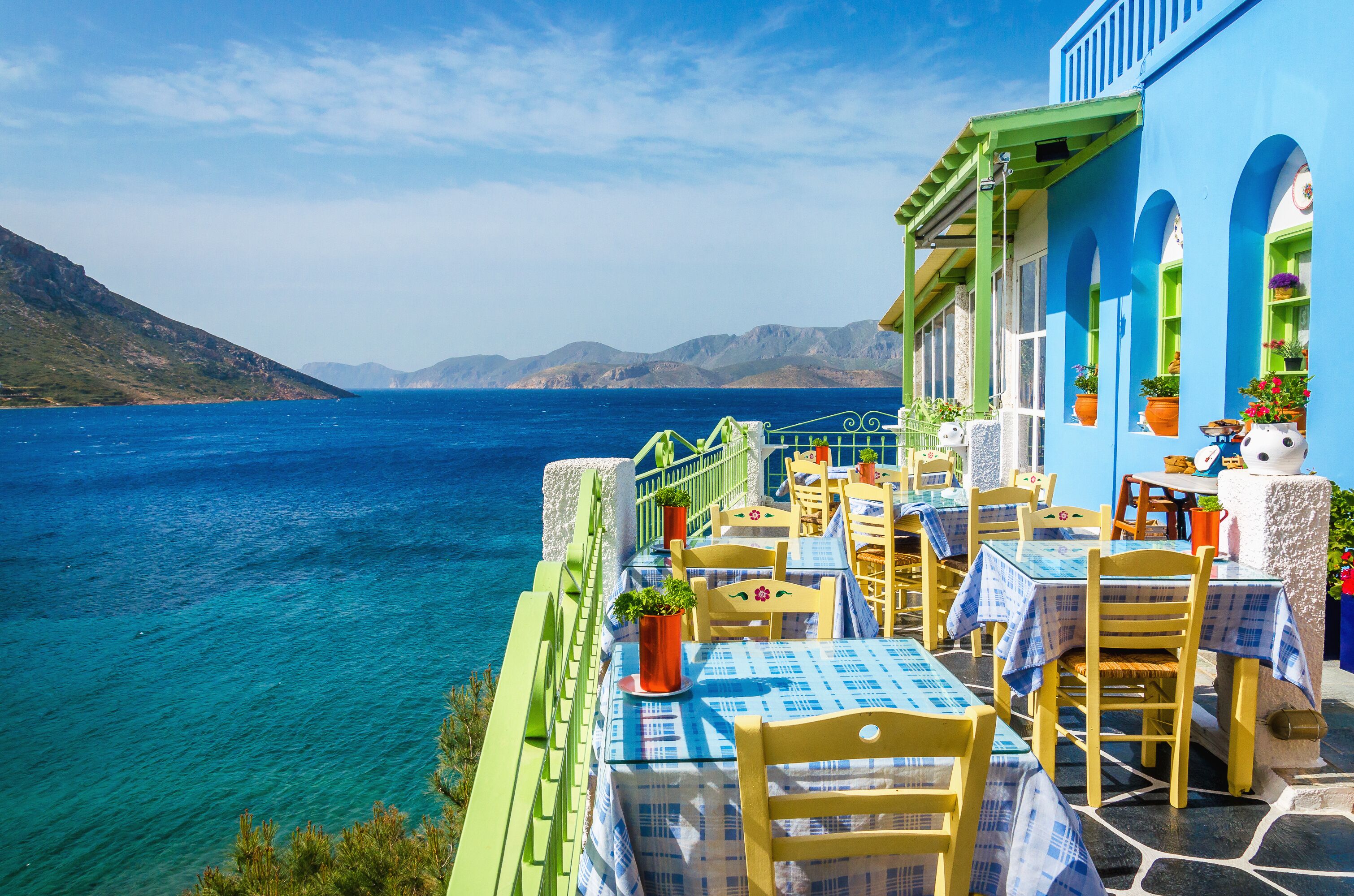 A seaside restaurant with blue and yellow tables and chairs, overlooking the sea and mountains.