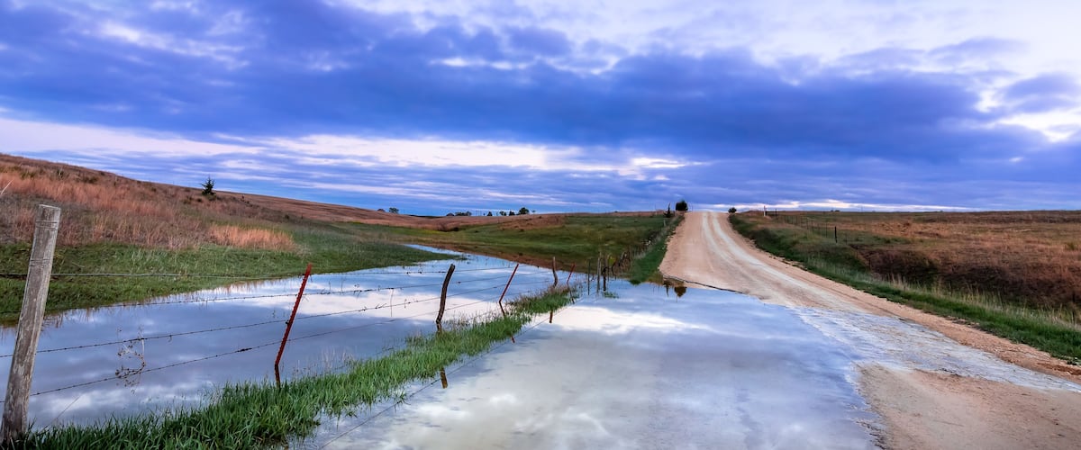 A rural landscape with a dirt road, a small water body, and a fence.