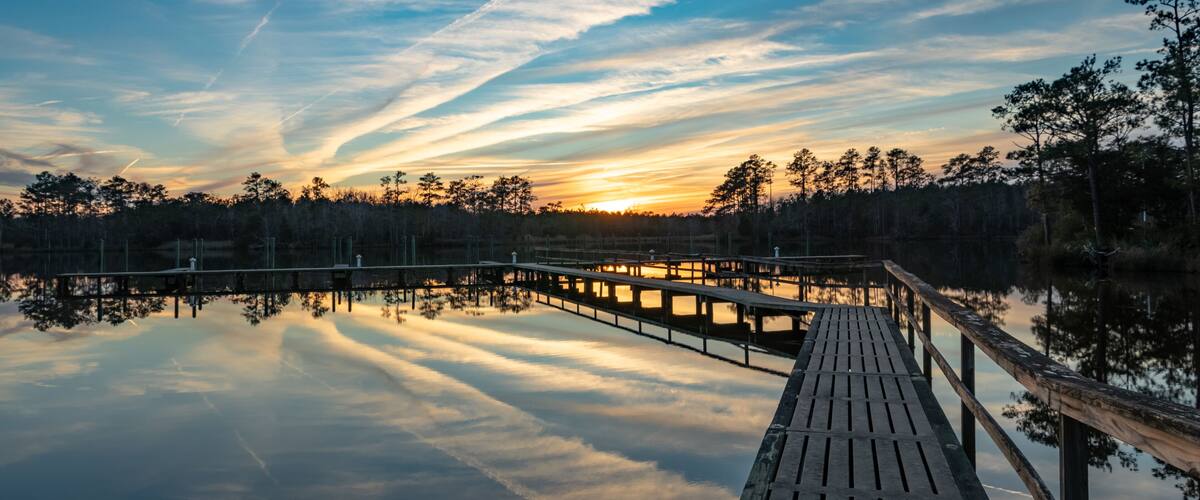 A wooden pier extending into a calm body of water, surrounded by trees and reflecting a sunset sky with wispy clouds.