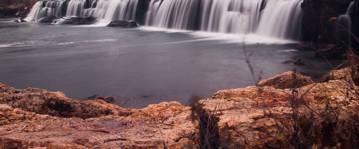 A multi-tiered waterfall cascading over rocky terrain with autumn-colored trees in the background.