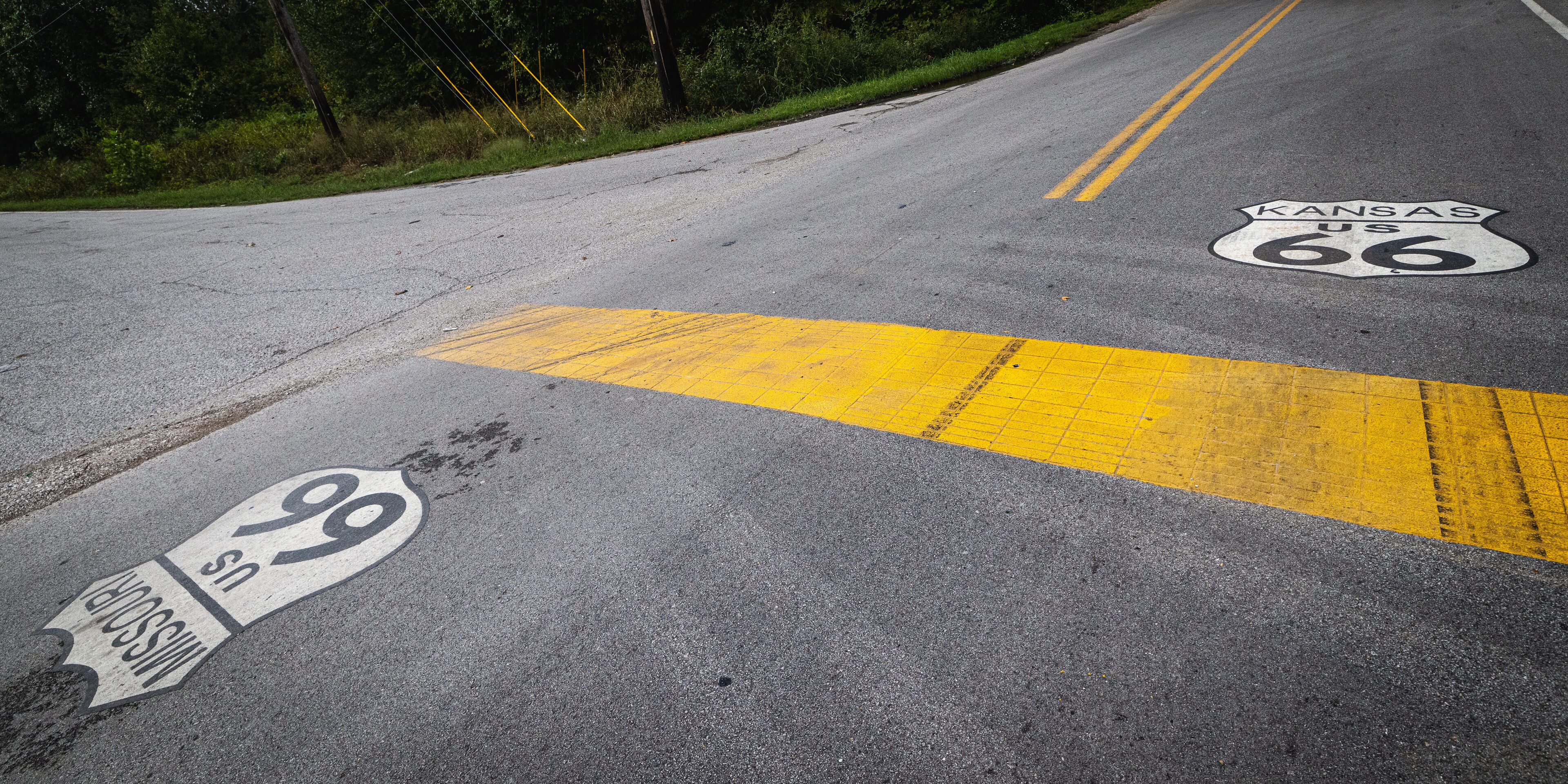 A high-angle view of the historic Route 66 road surface marking the border between Missouri and Kansas. A thick yellow line separates the painted shields on the asphalt pavement.