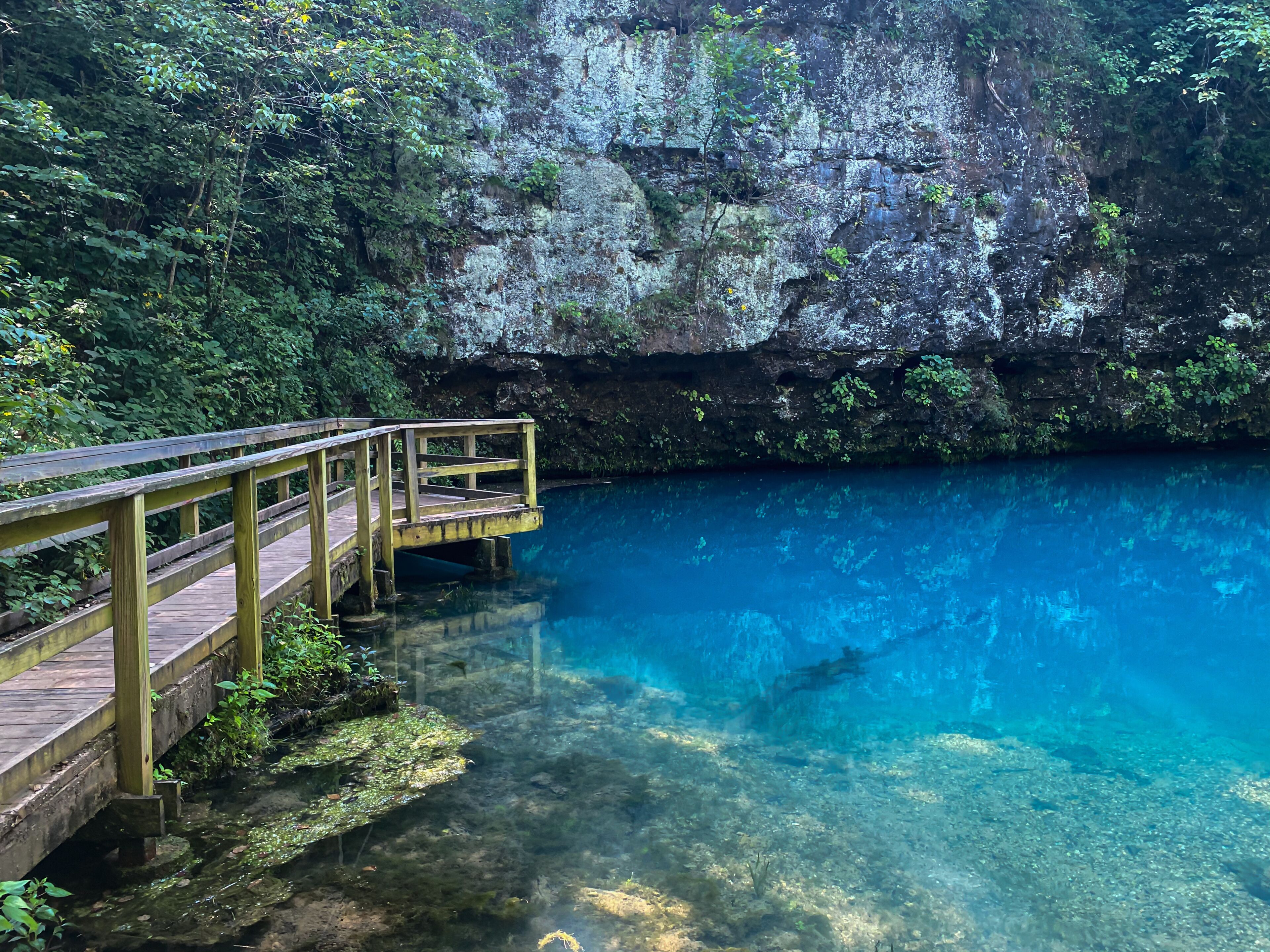 Uma ponte de madeira sobre uma piscina azul cristalina, com um penhasco rochoso ao fundo.