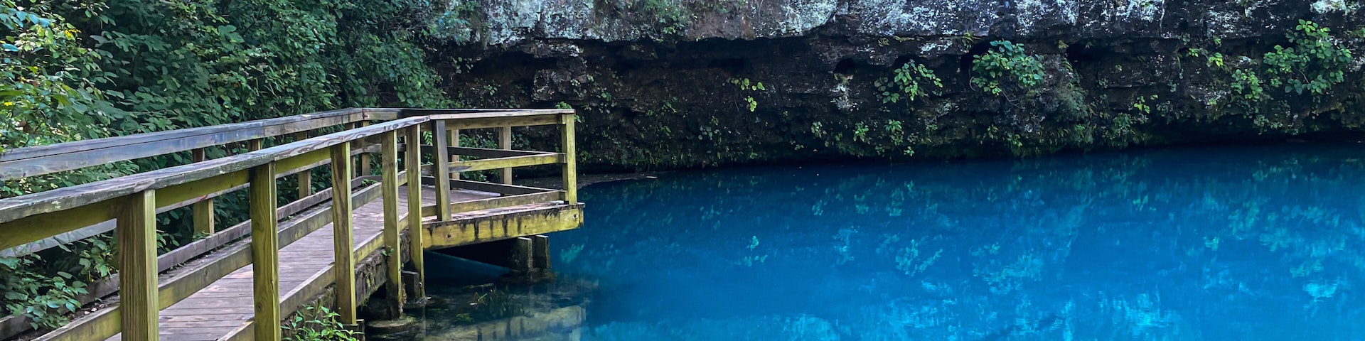 Uma ponte de madeira sobre uma piscina azul cristalina, com um penhasco rochoso ao fundo.