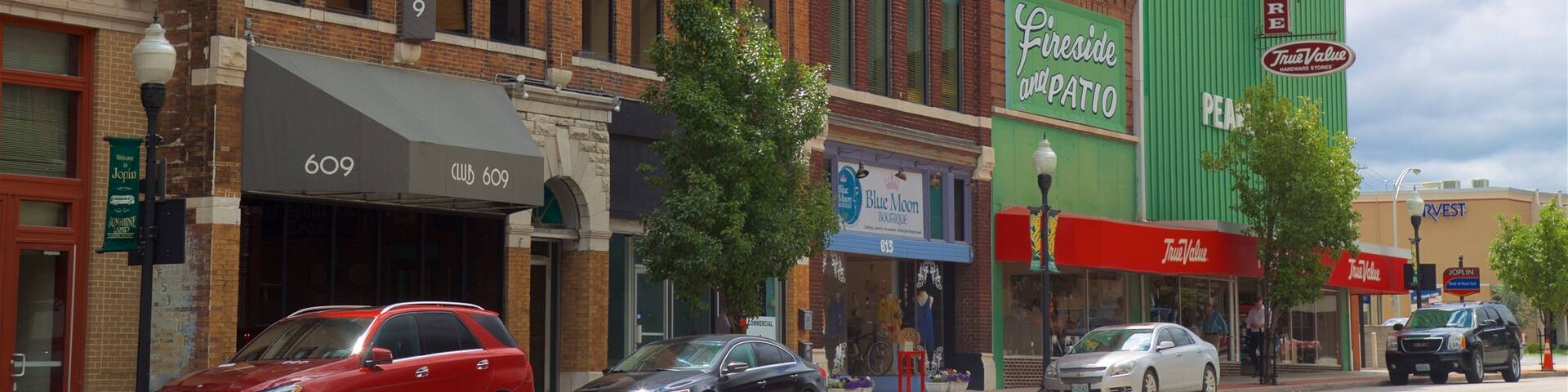A street with brick buildings, cars parked along the curb, and various storefronts including a hardware store and a boutique.