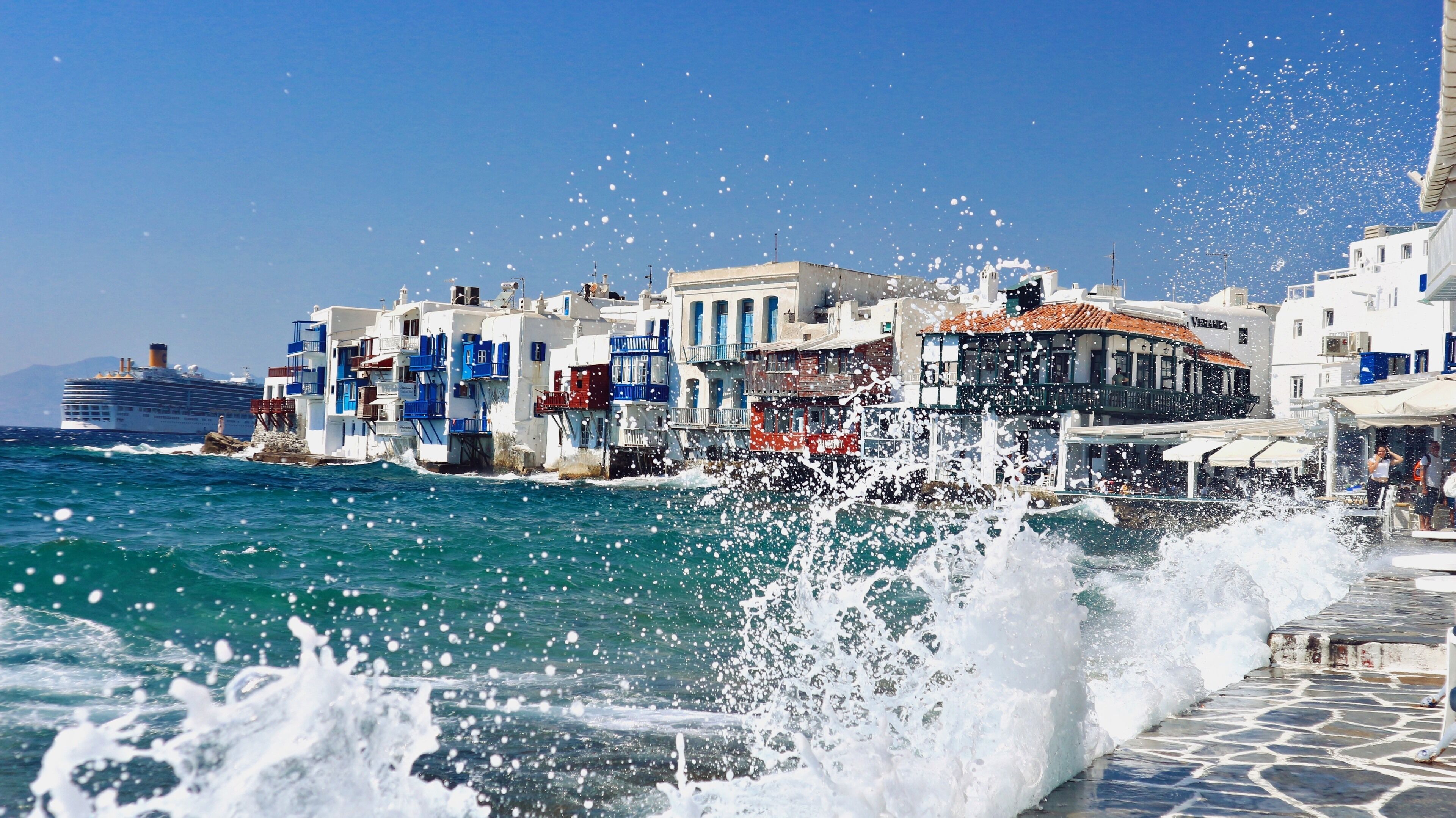 A coastal town with white buildings, a cruise ship, and waves crashing onto the shore.