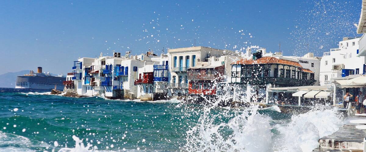 A coastal town with white buildings, a cruise ship, and waves crashing onto the shore.