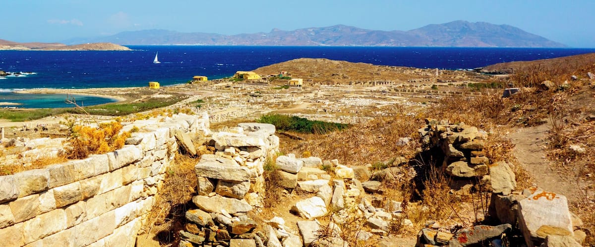 A coastal landscape with ruins, a sailboat, and distant mountains.