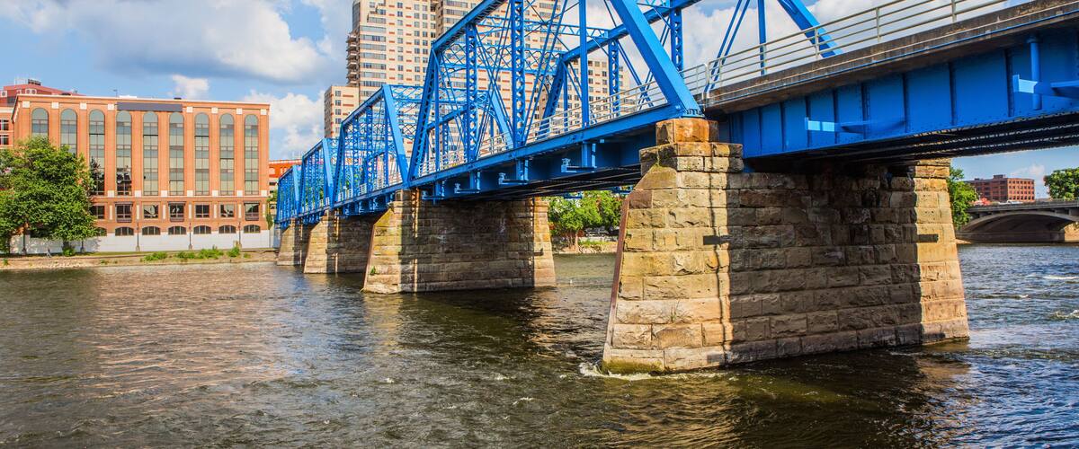 A blue steel bridge over a river with stone pillars, surrounded by buildings and trees.