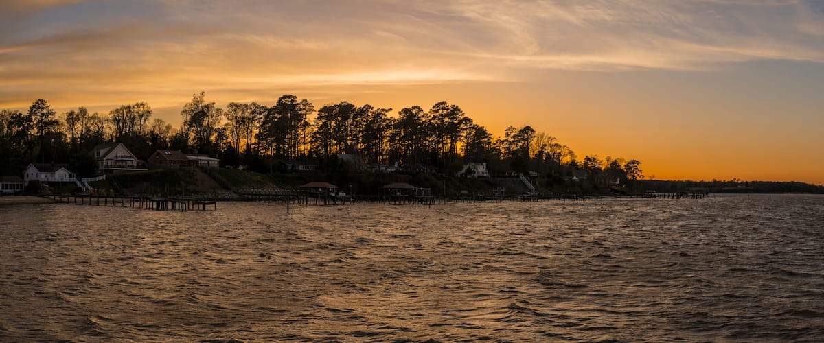 A sunset over a body of water with a row of houses and trees along the shore.