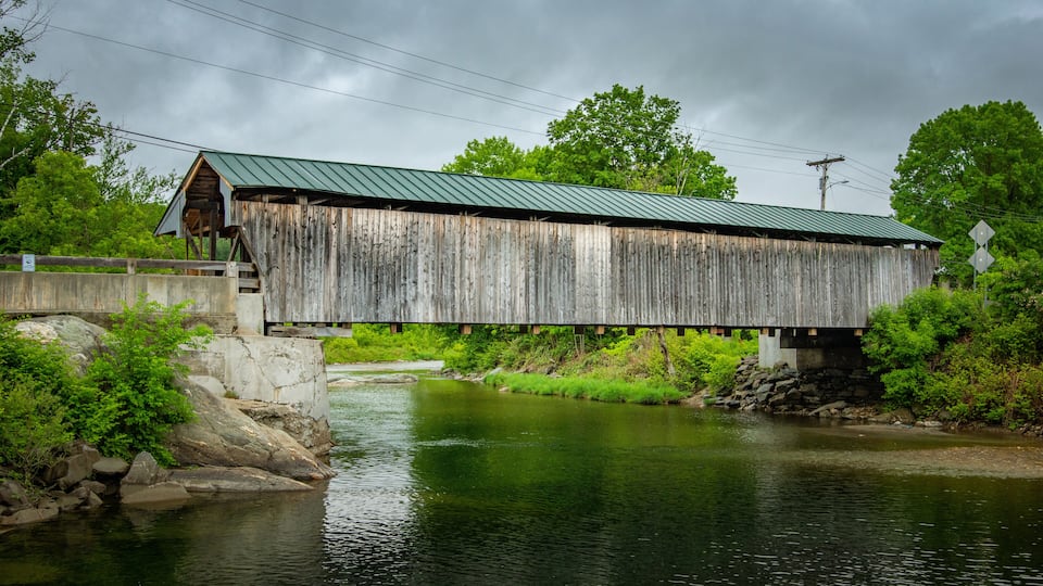 A wooden covered bridge over a river with a metal roof, surrounded by greenery and a cloudy sky.