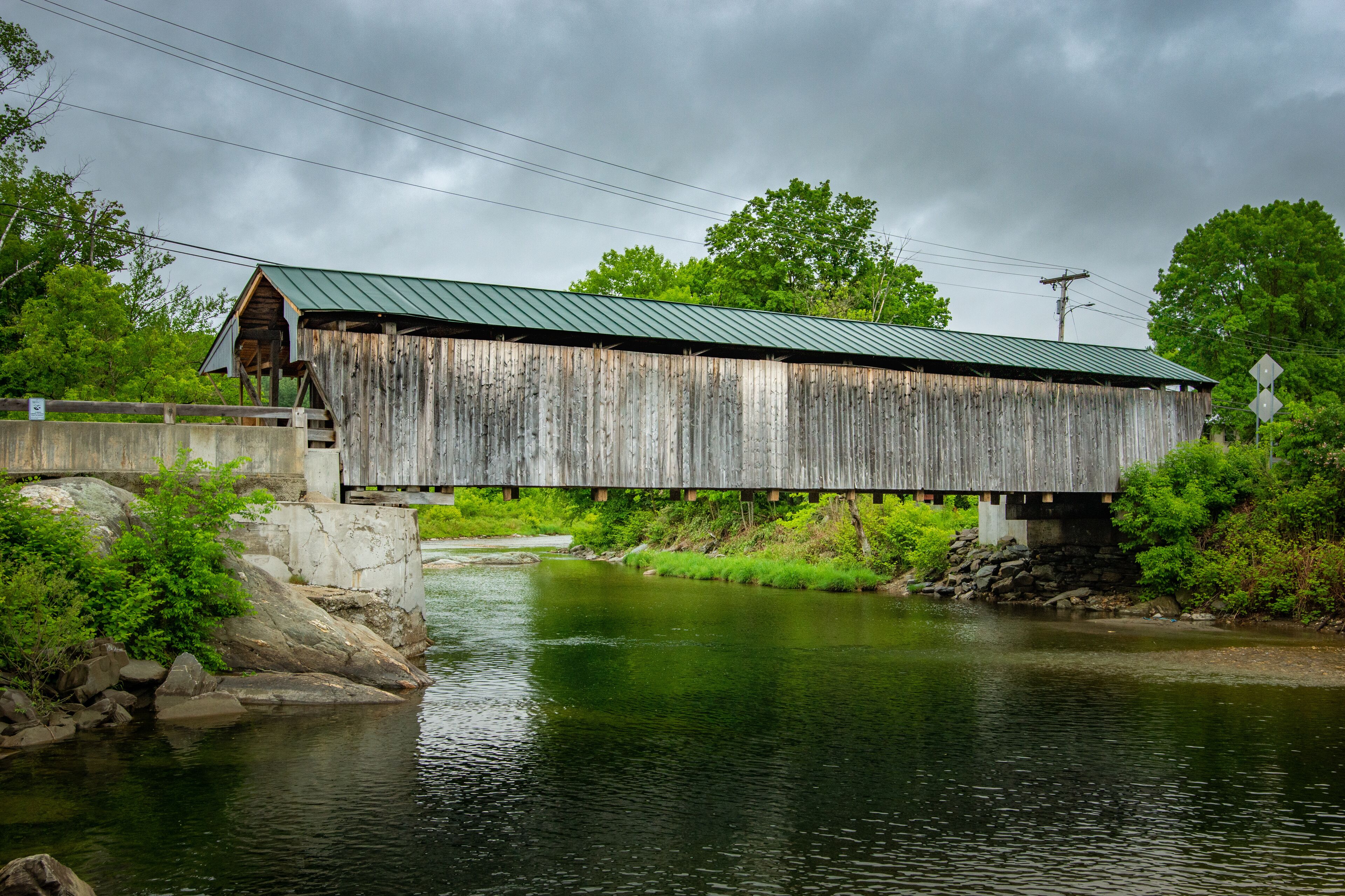 A wooden covered bridge over a river with a metal roof, surrounded by greenery and a cloudy sky.