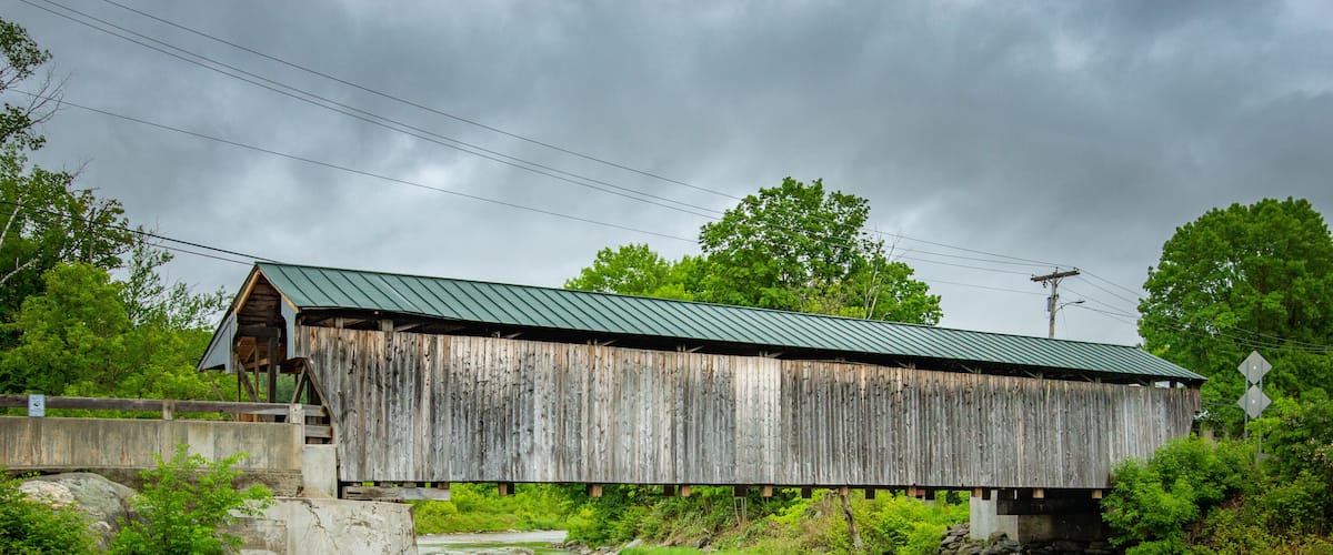 A wooden covered bridge over a river with a metal roof, surrounded by greenery and a cloudy sky.