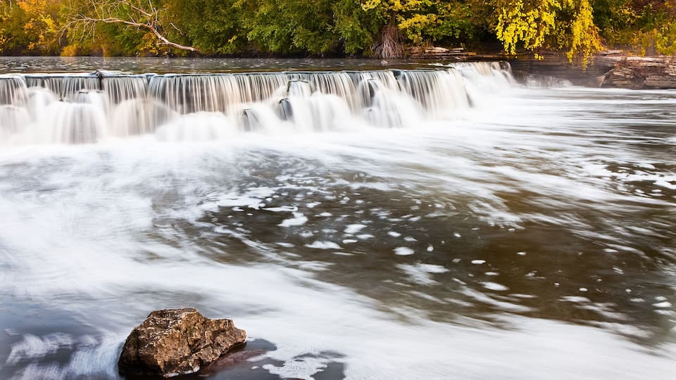 A waterfall with a rock in the foreground and autumn foliage in the background.