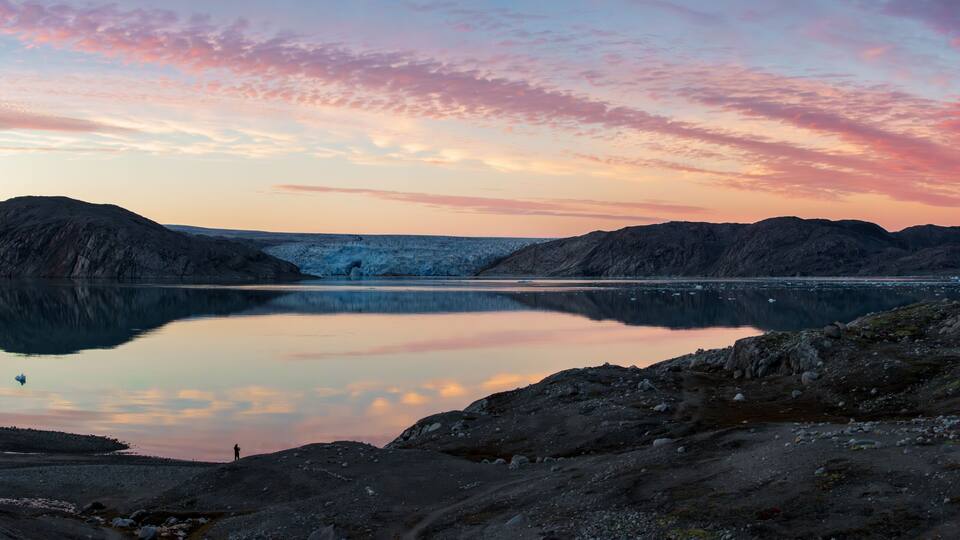 A calm lake with rocky terrain and mountains in the background during sunset.