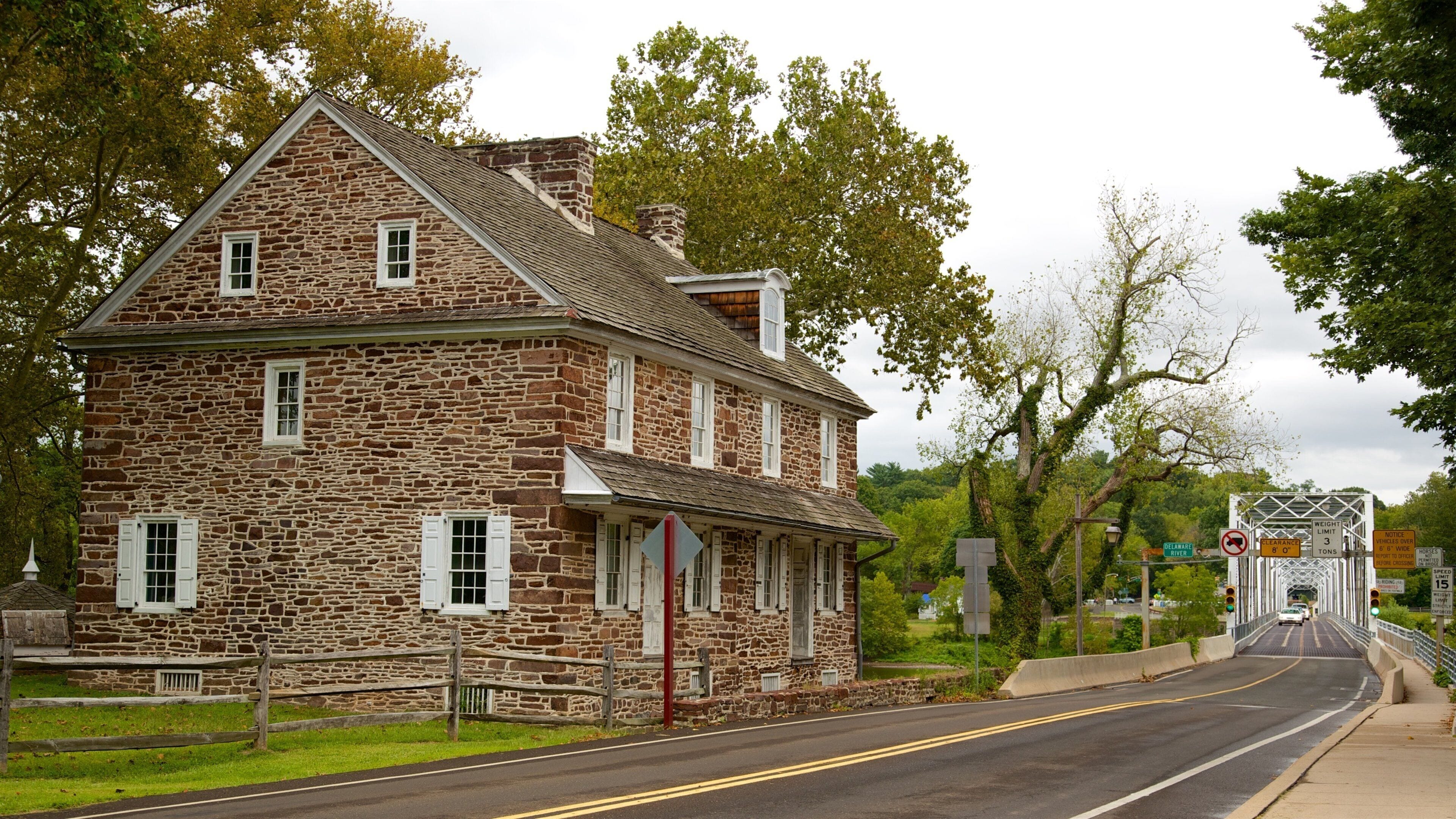 A stone house with white shutters and a red door, located beside a road with a bridge in the background.