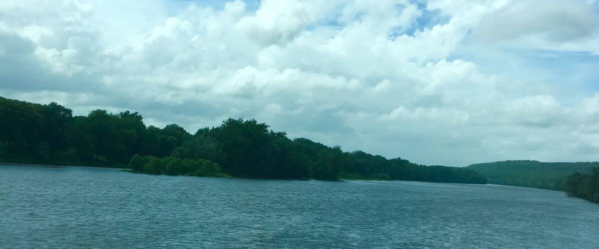 A calm river with a forested shoreline and a partly cloudy sky.
