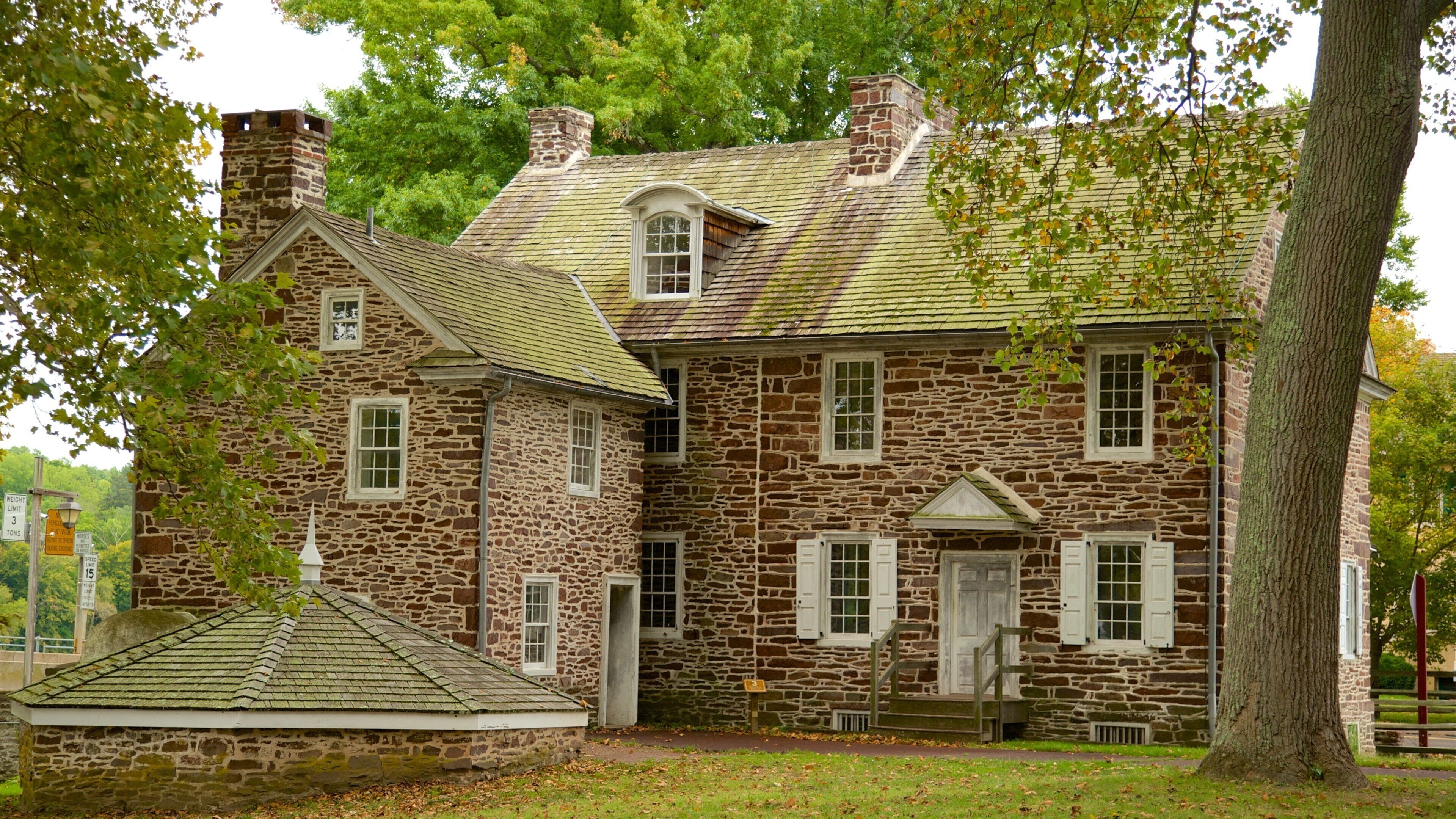A two-story stone house with a green roof, surrounded by trees and a small garden.
