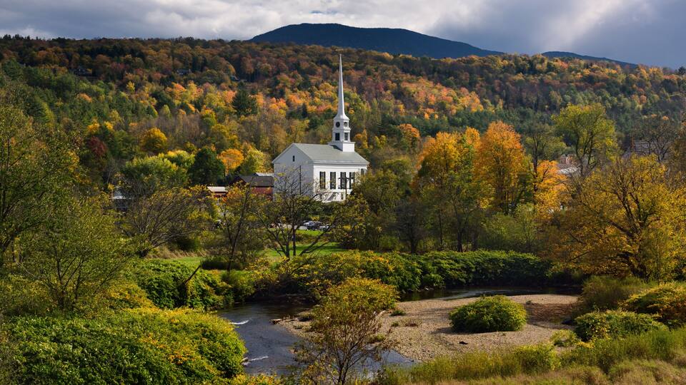A white church with a steeple surrounded by autumn foliage and a river.
