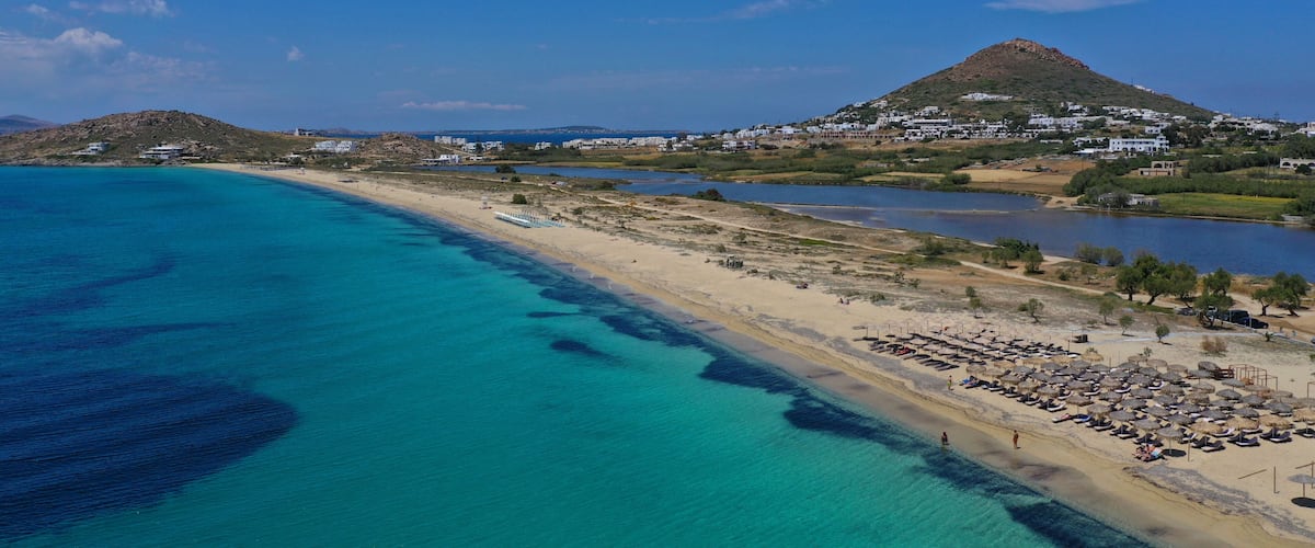 Een kustlandschap met een strand, helder turquoise water en een heuvel met gebouwen.