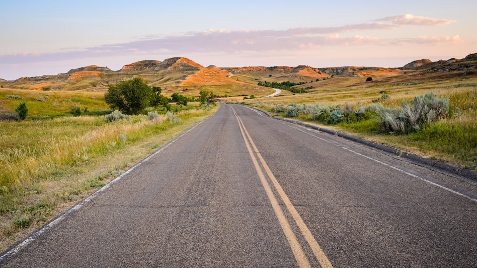 A two-lane road in a hilly, grassy landscape with sparse trees and distant hills.
