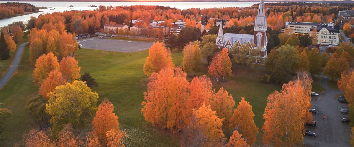 A town with autumn-colored trees, a church with a tall steeple, and a river in the background.