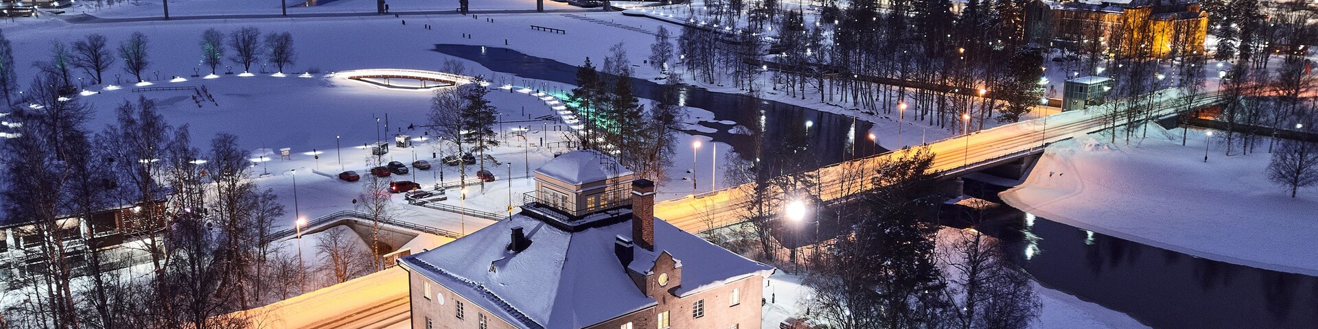 Un paisaje nevado con un río, un puente y edificios iluminados al anochecer.