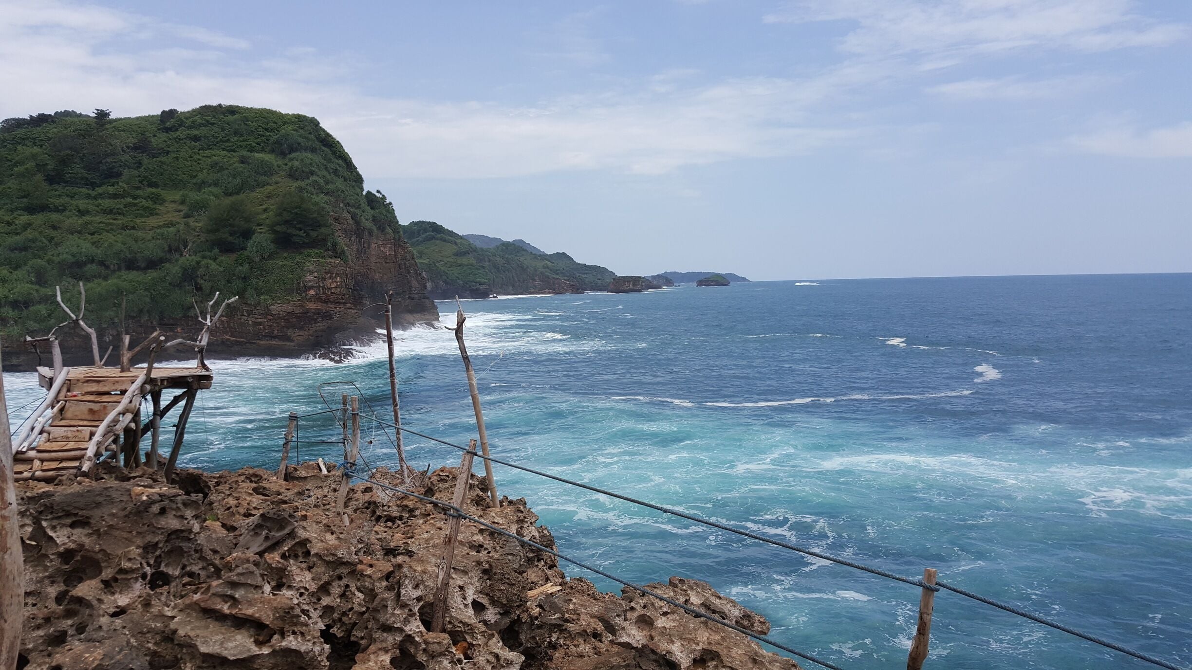 A coastal landscape with a wooden viewing platform overlooking the ocean, rocky cliffs, and a distant island.