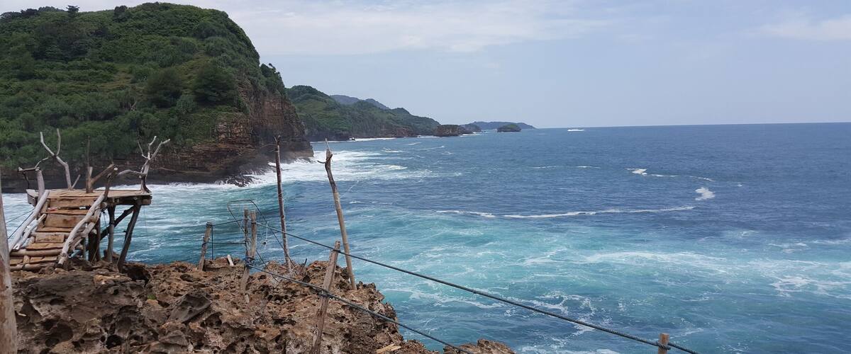 A coastal landscape with a wooden viewing platform overlooking the ocean, rocky cliffs, and a distant island.