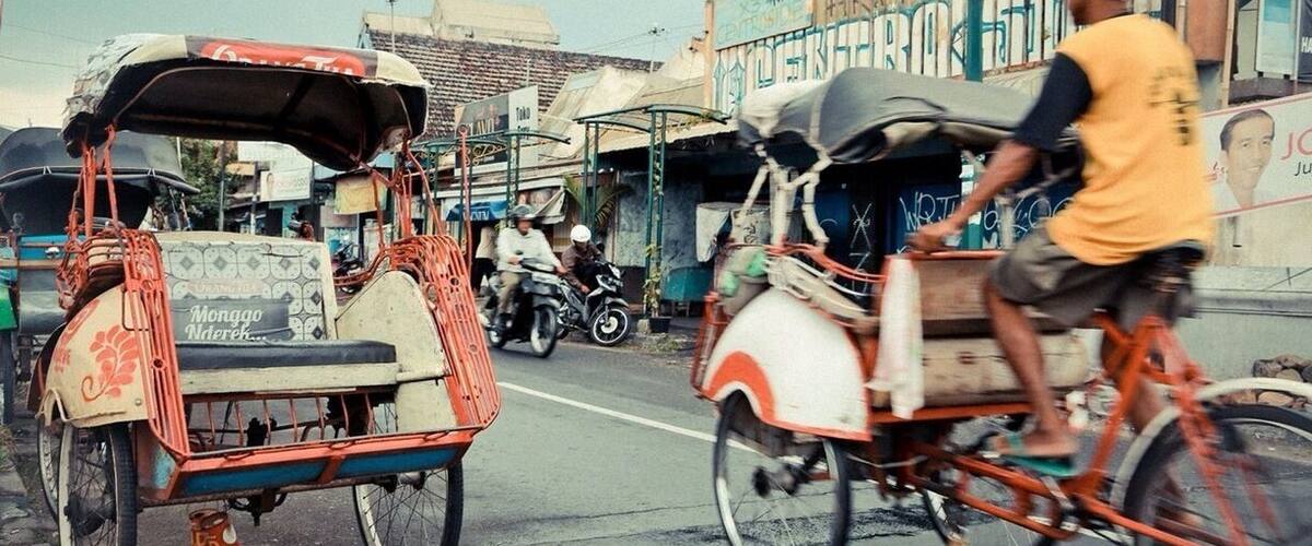 A street scene with a rickshaw and a motorcycle. There are buildings and a sign in the background.