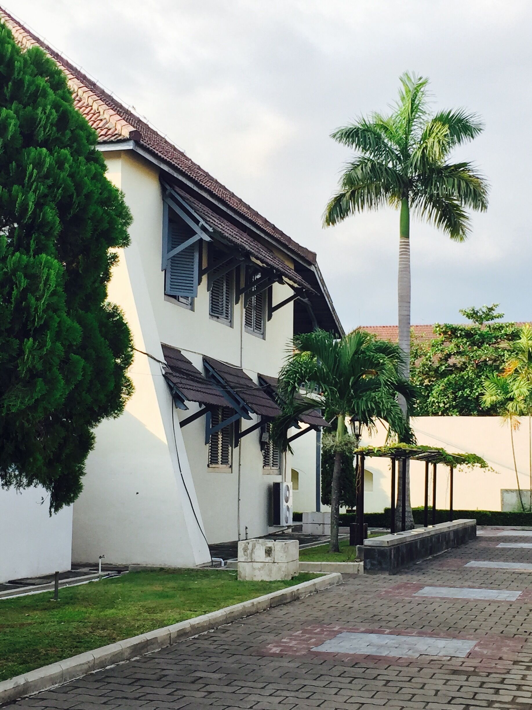 A white building with a tiled roof and shutters, surrounded by palm trees and a paved walkway.