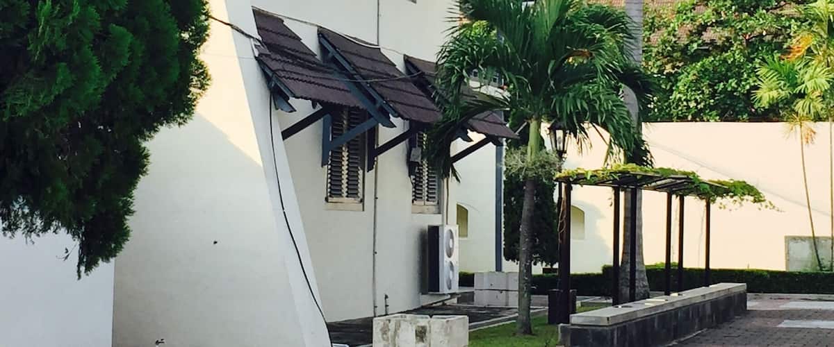 A white building with a tiled roof and shutters, surrounded by palm trees and a paved walkway.