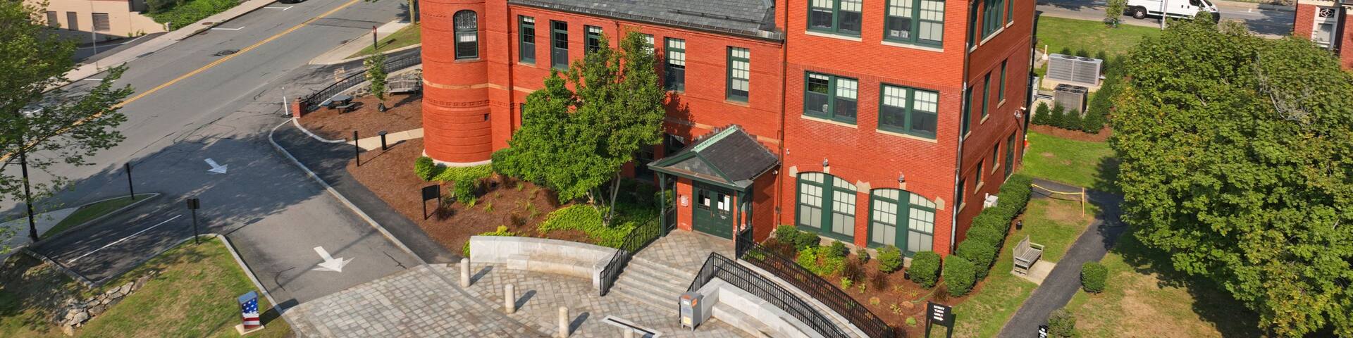 A red brick building with a tower, surrounded by greenery and a paved area with bollards.