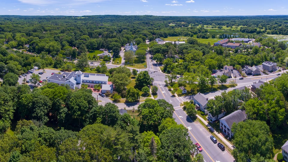 An aerial view of a town with a main road, residential houses, and surrounding greenery.