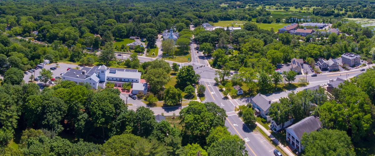 An aerial view of a town with a main road, residential houses, and surrounding greenery.
