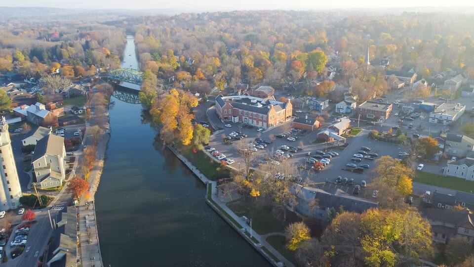 An aerial view of a town with a river, a bridge, and autumn-colored trees.