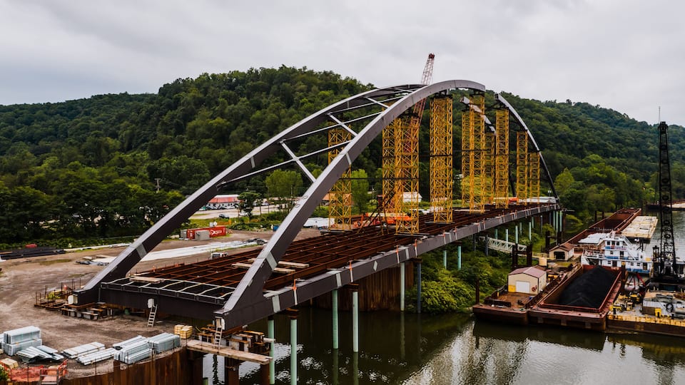 A large steel bridge with a curved arch spanning a river, surrounded by dense forest and industrial structures.